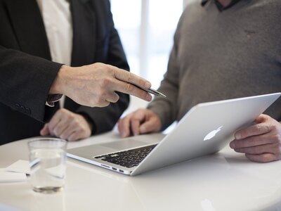 Laptop; im Hintergrund (angeschnitten) zwei diskutierende Personen